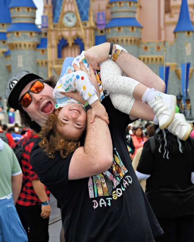 Craig holding his daughter upside down, both laughing, in front of Cinderella Castle at Magic Kingdom