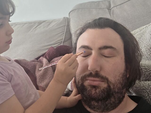 Craig on the sofa with his eyes closed while his daughter carefully applies eyeshadow